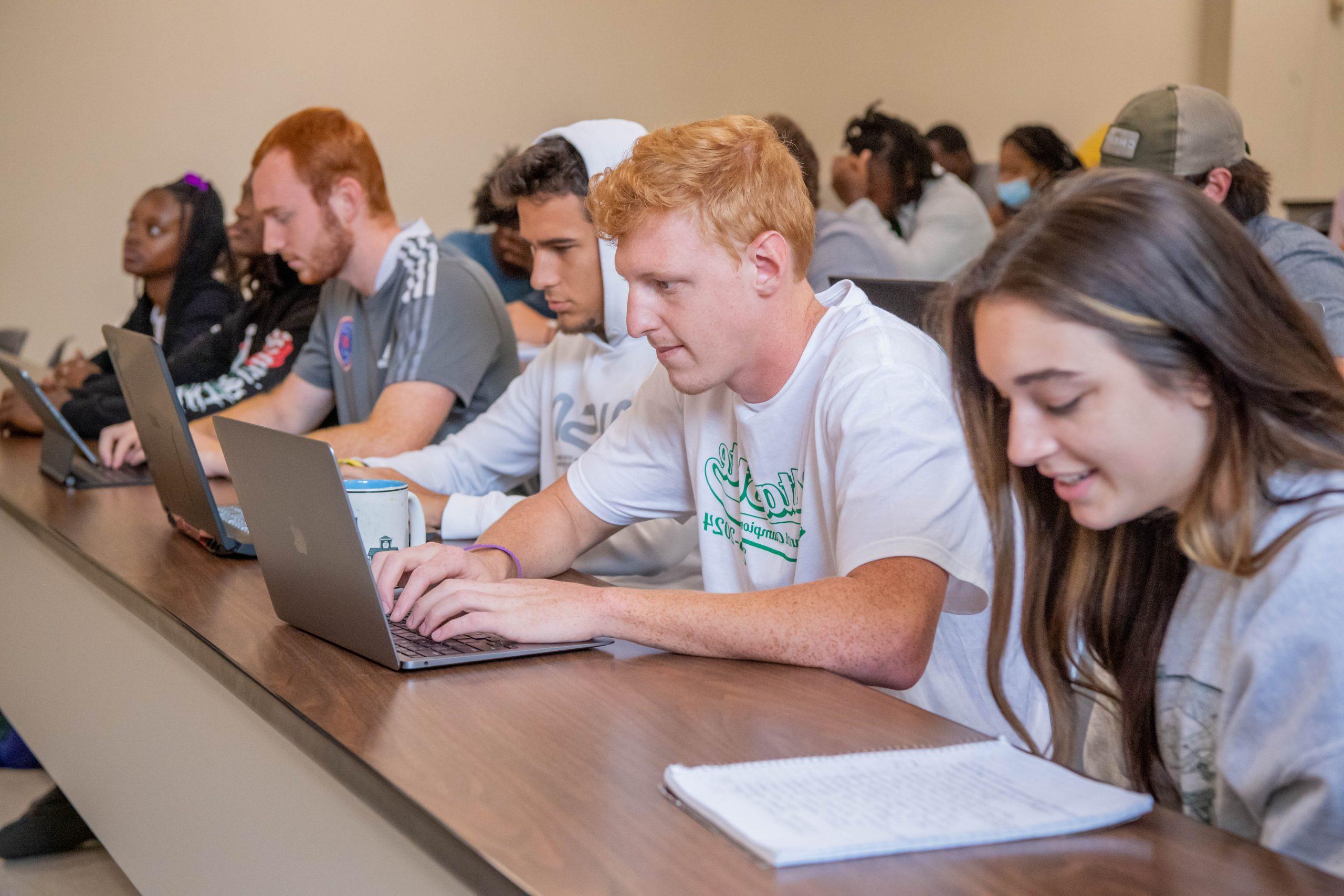 A front row of six students in a classroom taking notes on laptops and on paper.