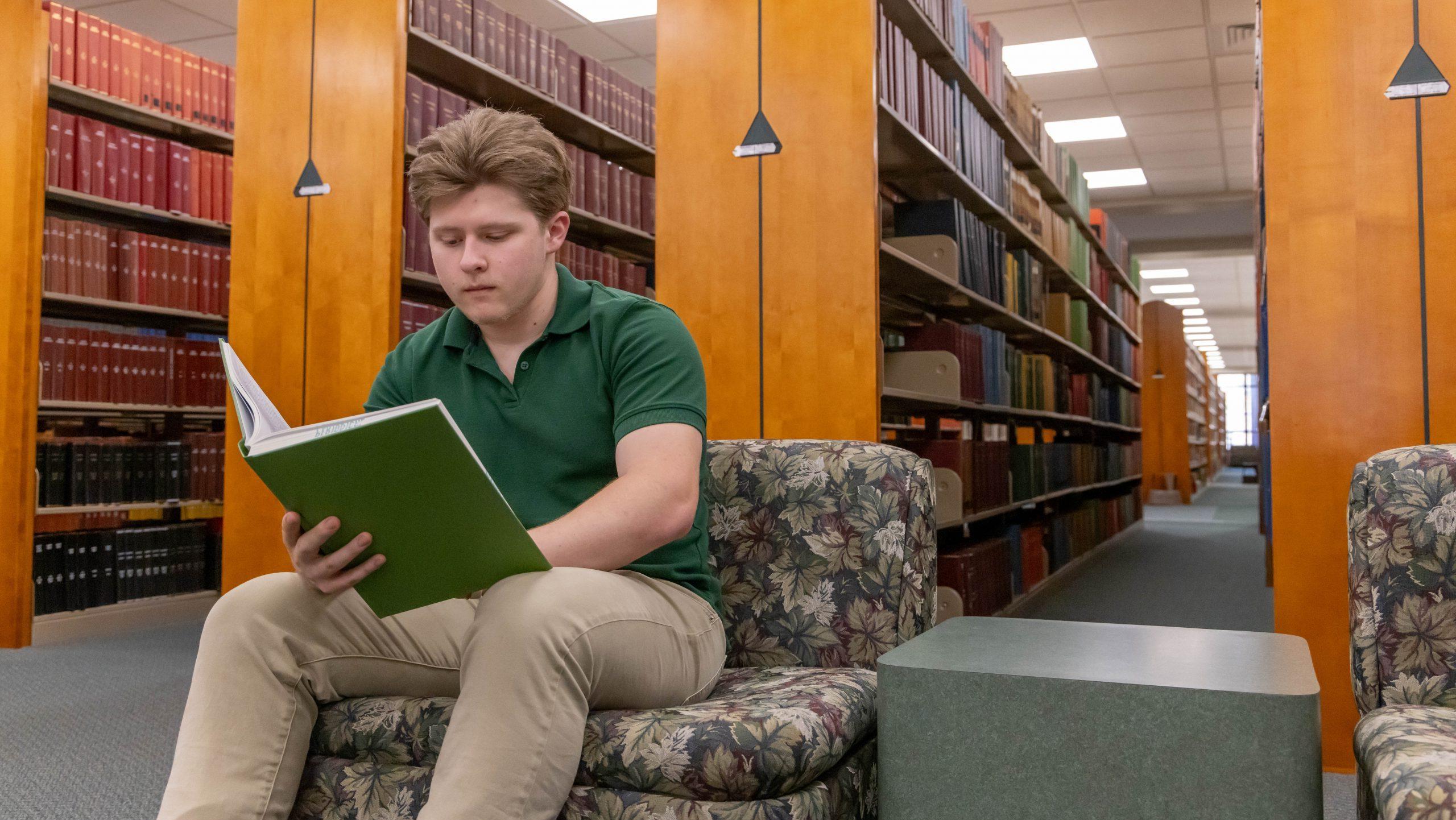 Student sitting near book shelves reading a book in the library.