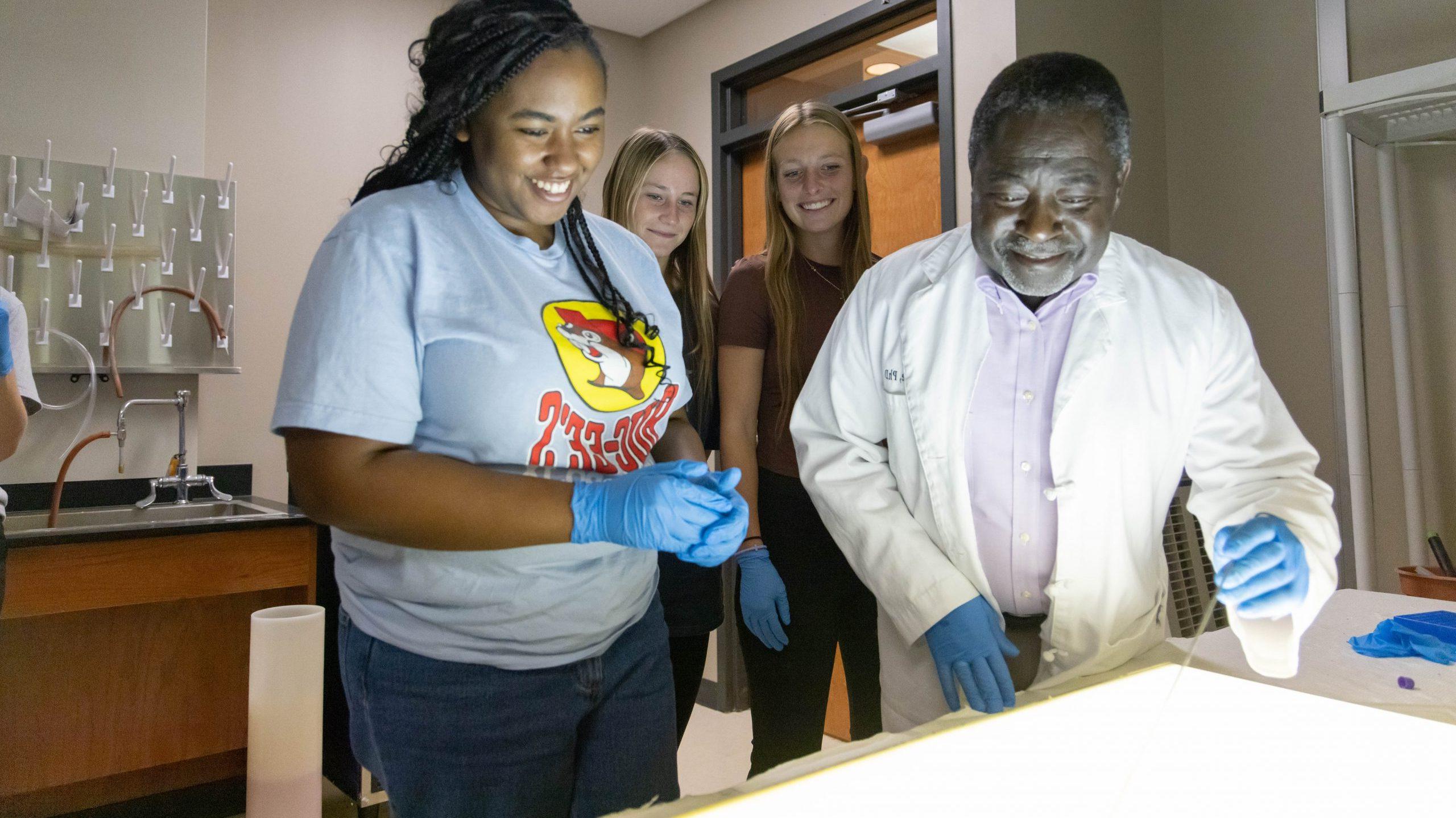 A professor and three students in a science lab smiling and engaging in a hands-on activity with the professor demonstrating something on a brightly lit surface.